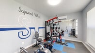 The Sagewood Villas Fitness Center is a dedicated workout space designed to provide residents with a convenient and functional exercise environment. The room features light gray walls prominently branded with the bold "SV" Sagewood Villas logo and lettering, giving the space a dynamic, athletic character. Interlocking foam floor tiles in gray and blue provide cushioning and protection underfoot, covering the majority of the exercise area.

The fitness center is equipped with a multi-station cable weight machine with red and black framing, a recumbent exercise bike, a rowing machine, and a treadmill, offering a well-rounded selection of both strength and cardio equipment. A full wall of framed mirrors on one side visually expands the space and allows users to monitor their form during workouts.

Climate comfort is ensured by a wall-mounted split-unit air conditioning system. Natural light filters in through a large window fitted with a roller shade, complementing the overhead flush-mount ceiling light. A keyed entry door with a glass panel provides secure, private access. A small waste bin and posted fitness center rules near the entrance complete the practical amenities. The textured ceiling and clean white baseboards add a polished, well-maintained finish to this efficiently arranged community fitness space.