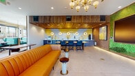 This thoughtfully designed amenity lounge delivers a striking mid-century modern aesthetic across a single, open floor. The space is anchored by a full-service bar and kitchen area, finished with bold yellow and white geometric tile work and illuminated by a series of round gold wall sconces. Warm wood-slat ceiling panels cascade over the bar zone, adding texture and visual depth, while a statement Sputnik chandelier with globe fixtures serves as a focal point overhead. A vibrant living moss wall frames a large-format display screen, creating a dynamic blend of natural and technological elements. Generous floor-to-ceiling windows line the perimeter, flooding the space with natural light and offering open views of the surrounding skyline. Seating is varied and plentiful, including a channeled amber leather sofa, upholstered banquette booths, and bistro-style tables paired with navy and gold accent chairs. A communal wood dining table anchors a secondary gathering area beside the kitchen, which is equipped with an oven, refrigeration, and sink. Light large-format tile flooring flows throughout, tying the zones together. Bold contemporary artwork and contrasting black-and-white graphic panels complete the curated, design-forward atmosphere.