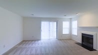 This single-floor space features a spacious, open living area designed with simplicity and functionality in mind. The room is finished with soft, patterned wall-to-wall carpeting that adds texture and comfort underfoot. Crisp white walls and a smooth white ceiling create a clean, neutral backdrop throughout.

Natural light is a defining characteristic of this space. A large sliding glass door fitted with vertical blinds serves as the focal point of the main wall, allowing generous amounts of daylight to enter while also providing direct access to an outdoor area. Flanking the opposite side of the room, two additional windows further brighten the interior, contributing to an airy and open atmosphere.

The layout is straightforward and versatile, offering ample room for a variety of furniture arrangements. Recessed ceiling vents are visible, indicating a central climate control system. The overall design is understated and move-in ready, making it well-suited for those looking to personalize the space to their own taste. The abundance of natural light and neutral finishes give the room a welcoming and comfortable feel.