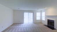 This single-floor space features a spacious, open living area designed with simplicity and functionality in mind. The room is finished with soft, patterned wall-to-wall carpeting that adds texture and comfort underfoot. Crisp white walls and a smooth white ceiling create a clean, neutral backdrop throughout.

Natural light is a defining characteristic of this space. A large sliding glass door fitted with vertical blinds serves as the focal point of the main wall, allowing generous amounts of daylight to enter while also providing direct access to an outdoor area. Flanking the opposite side of the room, two additional windows further brighten the interior, contributing to an airy and open atmosphere.

The layout is straightforward and versatile, offering ample room for a variety of furniture arrangements. Recessed ceiling vents are visible, indicating a central climate control system. The overall design is understated and move-in ready, making it well-suited for those looking to personalize the space to their own taste. The abundance of natural light and neutral finishes give the room a welcoming and comfortable feel.