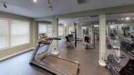 This dedicated fitness center is designed to support a full range of exercise routines within a single, open-plan floor. The space is finished with durable dark rubber flooring throughout, providing a practical and resilient surface suited for heavy equipment and high-impact workouts. Sage-green walls lend a calm, focused atmosphere to the environment, complemented by crisp white baseboards and structural columns that define zones within the open layout.

Cardio equipment is well represented, with multiple treadmills — including Matrix-brand units — as well as elliptical trainers and a stair climber positioned across the floor. A dedicated strength training area features cable weight machines and benches, with equipment mats anchoring each station. Floor-to-ceiling mirrors line a significant portion of the walls, visually expanding the space and allowing users to monitor their form during workouts.

Overhead track lighting illuminates the entire floor, ensuring consistent brightness across all exercise zones. Multiple windows fitted with white horizontal blinds allow natural light to supplement the artificial lighting while maintaining privacy. A built-in cubby storage unit is also present along one wall, providing convenient space for personal belongings. The overall ambiance is clean, functional, and professional, reflecting a thoughtfully outfitted fitness facility.