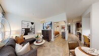 This well-designed single-floor residence offers a thoughtful layout that maximizes both function and style. The entry hallway, finished with light wood-tone flooring, leads directly into the heart of the unit. The galley-style kitchen is outfitted with dark wood cabinetry extending to the ceiling, stainless steel appliances including a French door refrigerator, a built-in microwave, dishwasher, and a tile backsplash complementing light quartz countertops. A compact built-in desk area with open shelving sits adjacent to the kitchen, ideal as a home workspace. The open-concept living room features large windows with horizontal blinds that invite natural light and offer views of surrounding greenery. The space accommodates a comfortable seating arrangement and is accented by contemporary artwork. The dining area is defined by track lighting overhead and a full-length floor mirror that visually expands the room, paired with a round table and seating for four beside a bright window. The bedroom, accessible directly from the living space, provides a private retreat. The bathroom is finished with large-format marble-style tile surrounding the soaking tub and shower combination, dark wood cabinetry, and neutral tile flooring, completing this clean and contemporary residence.
