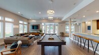 Located at 921 Trailwood Dr in Raleigh, NC, this single-floor clubhouse offers a well-appointed community gathering space designed with modern finishes and an open, social layout. The interior is anchored by rich hardwood-style flooring that flows throughout, complemented by neutral warm-toned walls and elevated ceilings that enhance the sense of openness.

The main gathering area is divided into distinct yet connected zones. A game area features a full-size billiard table positioned beneath stylish geometric pendant light fixtures, creating a focal point for social activity. Adjacent to this, a generously sized lounge area is furnished with mid-century modern seating arrangements, including sofas and accent chairs grouped around a marble-top coffee table atop a bold, patterned area rug. Large windows with transom panels line the walls, flooding the space with abundant natural light and offering views of the surrounding outdoor landscape. French doors provide direct access to an outdoor pergola area.

The kitchen is a standout feature, showcasing expansive granite countertops, dark wood cabinetry with a decorative tile backsplash, stainless steel appliances, and a bar-height seating counter with pendant lighting above. A wall-mounted television and additional seating areas complete this versatile, amenity-rich space.