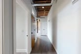 Located at 119 Hotel St in Utica, NY, this one-bedroom, one-bathroom loft masterfully blends industrial character with contemporary design. The open-concept kitchen and living area serves as the heart of the space, showcasing soaring exposed wood beam ceilings, dramatic exposed brick walls, and large black-framed windows that flood the interior with natural light and offer views of the surrounding cityscape. A large opening in the brick wall further enhances the connection to the outdoors. The kitchen is well-appointed with stainless steel appliances, white upper cabinetry, a subway tile backsplash, and a central island with a sleek countertop and slate-blue cabinetry, pendant lighting overhead adds a refined touch. Hardwood flooring runs throughout the main living areas, complementing the raw industrial aesthetic. The hallways feature exposed ductwork and structural ceiling joists, reinforcing the loft's character. The bedroom offers an intimate retreat with a natural wood floor, an exposed brick accent wall, and a window providing natural light. A dedicated closet provides practical storage. The bathroom is finished with classic white hexagonal tile flooring, a glass-enclosed shower, a vanity with dark cabinetry, and an exposed brick accent wall that ties back to the overall design theme.