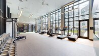 This contemporary fitness center is thoughtfully designed to accommodate a wide range of workout styles within a spacious, single-level layout. The facility is divided into distinct functional zones, each finished with appropriate flooring—durable rubber matting in the cardio and strength areas and light hardwood in the stretching and studio section.

The main cardio area runs along an expansive wall of floor-to-ceiling windows framed in dark steel, flooding the space with natural light and offering direct views of the outdoor courtyard. A row of treadmills, ellipticals, and stair climbers lines this windowed wall, all equipped with individual screens. Ceiling fans and suspended linear lighting maintain comfort and visibility throughout.

A dedicated strength training zone features a slatted functional training wall mounted with resistance bands, cables, and accessories, alongside a multi-use cable machine and weight bench. Free weights, kettlebells, medicine balls, and stability balls are neatly organized on racks in an adjacent area.

The studio section offers a mirrored wall spanning its full length, a wall-mounted television with a teal accent surround, and hardwood flooring suited for yoga, stretching, or personal training. Black-framed glass doors and partitions connect the zones while maintaining visual openness throughout the entire facility.