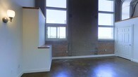 This two-story space showcases an industrial loft aesthetic defined by its dramatic vertical scale and raw architectural character. The interior features exposed brick walls that add warmth and texture, contrasting with polished concrete floors that reflect light and enhance the sense of openness throughout. Towering ceilings accommodate a generous arrangement of large, multi-pane windows spanning both levels, allowing abundant natural light to pour in while framing views of the surrounding treetops and skyline. A partial upper-level partition with a warm wood-capped ledge is visible, suggesting a mezzanine or second-floor overlook that adds depth and dimension to the layout. Structural concrete columns punctuate the space, reinforcing its industrial origins while contributing to the overall architectural character. The combination of natural materials—brick, wood, and concrete—creates a layered, textural ambiance that balances ruggedness with refinement. The open floor plan and soaring heights make this space highly versatile, lending itself well to a variety of uses, from creative studios to live-work environments.