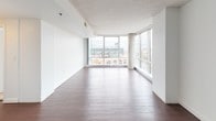 This single-floor residence offers a well-organized layout with distinct living, sleeping, and utility zones. The open living and dining area is a standout feature, finished with rich dark hardwood flooring that extends throughout the main space. Floor-to-ceiling windows line the exterior wall, flooding the interior with natural light and providing impressive views of the surrounding urban landscape, including prominent stadium structures visible from multiple vantage points.

The kitchen is designed in a U-shaped configuration with warm medium-toned wood cabinetry, granite countertops featuring earthy tones, a deep undermount sink, and a full suite of black appliances including a refrigerator, stove, and dishwasher. Overhead flush-mount lighting provides consistent illumination throughout the space.

The bedrooms are carpeted and bright, each benefiting from large corner or wraparound windows that frame city views. One bedroom includes access to a dedicated walk-in closet with a full-width hanging rod, offering generous storage. The bathroom is finished with small-format tile flooring, a white vanity with ample drawer storage, a mirrored medicine cabinet, and a full soaking tub with shower combination. The overall aesthetic is clean and contemporary throughout.