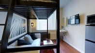 This compact yet thoughtfully designed single-floor space makes a strong visual statement through its use of contrasting black and white elements. The centerpiece of the living area is a dramatic black canopy frame with slatted detailing and a zebra-print accent above, creating a defined lounge zone within the open layout. Beneath the canopy, a white upholstered sofa is arranged with black and patterned throw pillows, complemented by a low white coffee table that anchors the seating area. Rich hardwood flooring in a warm reddish-brown tone runs throughout, adding depth and warmth to the otherwise high-contrast aesthetic. Natural light filters generously through a set of large windows fitted with vertical blinds, brightening the interior. A bold zebra-print framed artwork adorns one wall, reinforcing the space's daring design sensibility. Adjacent to the living area, a wall-mounted sink and vanity mirror introduce a utilitarian element, while a wall-mounted flat-screen television adds a modern, functional touch. The overall ambiance is sleek and eclectic, blending urban edge with comfortable living in a highly curated, single-room footprint.