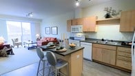 This well-designed single-floor space offers an open-concept layout that effortlessly blends the kitchen, dining, and living areas into one cohesive and functional environment. The kitchen is outfitted with light maple cabinetry, dark countertops, a full suite of white appliances including a range, microwave, and dishwasher, and a undermount sink. A central kitchen island with a dark countertop serves double duty as a casual dining spot, complemented by modern bar-height stools — making it an ideal space for both meal prep and entertaining. Track lighting overhead keeps the kitchen bright and practical. The open living area flows naturally from the kitchen, featuring light hardwood-style flooring and neutral wall tones that create a clean, airy atmosphere. A comfortable seating area anchors the living space, with framed artwork adding a personal touch to the walls. Large windows allow natural light to pour in, enhancing the overall sense of openness. The cohesive design, warm finishes, and smart use of space make this a highly livable and versatile floor plan suited for everyday comfort and social gatherings alike.