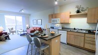 This well-designed single-floor space offers an open-concept layout that effortlessly blends the kitchen, dining, and living areas into one cohesive and functional environment. The kitchen is outfitted with light maple cabinetry, dark countertops, a full suite of white appliances including a range, microwave, and dishwasher, and a undermount sink. A central kitchen island with a dark countertop serves double duty as a casual dining spot, complemented by modern bar-height stools — making it an ideal space for both meal prep and entertaining. Track lighting overhead keeps the kitchen bright and practical. The open living area flows naturally from the kitchen, featuring light hardwood-style flooring and neutral wall tones that create a clean, airy atmosphere. A comfortable seating area anchors the living space, with framed artwork adding a personal touch to the walls. Large windows allow natural light to pour in, enhancing the overall sense of openness. The cohesive design, warm finishes, and smart use of space make this a highly livable and versatile floor plan suited for everyday comfort and social gatherings alike.