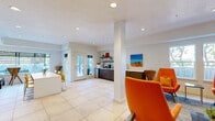 Located at 3808 S West Temple in South Salt Lake, Utah, this single-floor community amenity space is designed to foster comfort, creativity, and connection. The open-concept layout is anchored by large-format light tile flooring that runs seamlessly throughout, creating a bright and cohesive aesthetic.

The main lounge area is generously proportioned, featuring plush gray sectional sofas accented with vibrant red and orange throw pillows, a bold multicolored area rug, and a large wall-mounted flat-screen TV set atop a distinctive textured wood console. Tray ceilings with recessed lighting add architectural depth, while multiple windows invite abundant natural light and views of the outdoor patio area.

A secondary seating nook highlights bold yellow accent walls paired with vivid orange armchairs, adding an energetic pop of color. This area transitions toward a game room space equipped with a foosball table, offering a lively recreational option.

The kitchenette area is thoughtfully appointed with dark wood cabinetry, open floating shelves, a coffee station, and a compact refrigerator. Yellow bar-height chairs line a clean white counter near glass French doors that open directly to an outdoor patio, blurring the line between indoor and outdoor living.

Curated artwork, indoor plants, and statement décor pieces like an oversized orange ceramic vase complete the space, giving it a polished yet welcoming character throughout.