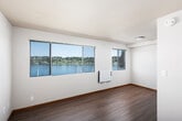 This single-floor space is thoughtfully designed to maximize comfort and natural light, with large windows throughout that frame stunning waterfront and hillside views. The main living area is open and airy, featuring warm wood-style laminate flooring, neutral-toned walls, and a textured ceiling. Wall-mounted heating units provide efficient climate control in multiple rooms.

The kitchen is equipped with modern stainless steel appliances, including an electric range, over-the-range microwave, and a full-size refrigerator. Dark wood cabinetry with brushed hardware pairs with light quartz countertops to create a clean, contemporary aesthetic.

A secondary room, likely a bedroom or den, also benefits from waterfront-facing windows with blinds for privacy, along with its own wall heater and wood-style flooring consistent with the rest of the unit. The bathroom features a full tub-shower combination with brushed nickel fixtures and a handheld showerhead, complemented by clean white finishes and a warm wood-toned door. The overall layout, as seen in the dollhouse view, is compact yet efficiently organized, with a clear separation between the living, kitchen, bathroom, and bedroom areas.