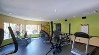This fitness center is a well-equipped, single-floor exercise space designed to support a range of workout routines. The room is immediately recognizable by its bold two-tone wall treatment, featuring a bright lime green upper section paired with a neutral cream lower portion, divided by a dark horizontal accent stripe. This color scheme gives the space an energetic, motivating atmosphere.

The facility is outfitted with a solid selection of equipment. Cardio options include a StairMaster step climber, an elliptical cross-trainer, stationary bikes, and a treadmill, all positioned to make efficient use of the available floor space. For strength training, a multi-station cable machine provides users with options for a variety of resistance exercises, complemented by a weight bench and additional strength apparatus.

A textured white ceiling with recessed lighting provides even illumination throughout the space, while windows along one wall allow natural light to filter in. A posted "Fitness Area Rules" sign keeps usage guidelines clearly visible to all members. The dark rubberized flooring is both practical and durable, ideal for high-traffic exercise use.