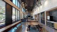 Located at 430 Jackson St in Oakland, CA, this single-floor amenity space combines industrial-chic aesthetics with modern comfort. The expansive main hall is defined by soaring ceilings with exposed ductwork and dramatic cluster pendant lighting featuring globe-shaped bulbs, casting a warm, inviting glow over polished concrete floors. Floor-to-ceiling windows along one wall flood the interior with natural light and offer views of the tree-lined streetscape outside.

The space is organized into distinct activity zones. A recreational area at the center is anchored by a full-size ping-pong table and a shuffleboard table, ideal for casual social gatherings. Adjacent to this, a lounge seating area features plush, high-backed velvet armchairs and sofas arranged over a colorful area rug, creating a comfortable retreat. A wall-mounted fireplace and flat-screen TV form a focal point for relaxation in this zone.

A dedicated coworking area offers light wood workstations and tables with ample room for productivity, situated near a kitchenette outfitted with wood cabinetry, a sink, and under-counter appliances. Warm wood paneling and white brick walls add texture and contrast throughout, while a semi-private room with black-framed glass partitions provides a quieter workspace option. The overall design blends raw industrial elements with refined furnishings for a dynamic, multi-functional environment.