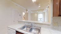 This two-floor space showcases a thoughtfully designed interior that blends classic architectural charm with functional living. The open-concept layout connects the kitchen and living areas seamlessly, creating a sense of spaciousness and flow throughout the main level.

The kitchen is equipped with a double stainless steel sink set into a generous marble-tiled countertop, offering ample workspace for meal preparation. A raised breakfast bar separates the kitchen from the adjoining living area while maintaining visual continuity between the two spaces.

The living area is a standout, featuring decorative columns that add a touch of classical elegance to the room. Exposed beam detailing along the ceiling adds architectural character, while track lighting throughout ensures the space is well-illuminated. An arched window serves as a focal point, allowing natural light to pour into the room and creating an airy, welcoming atmosphere. White walls and neutral tones throughout contribute to a bright and clean aesthetic.

Crown molding and carefully considered interior details elevate the overall design, reflecting a commitment to quality craftsmanship. The two-floor layout provides additional living or functional space beyond the main level, making this a versatile and well-appointed property suitable for a variety of uses.