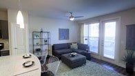 This single-floor space features an open-concept layout that seamlessly integrates the living and dining areas into one cohesive environment. The living area is anchored by a large gray sectional sofa accented with decorative pillows, paired with a matching ottoman that doubles as a coffee table. A plush, light-toned area rug defines the seating zone and adds texture to the dark hardwood flooring beneath. A ceiling fan with a built-in light fixture is centrally mounted overhead, providing both airflow and ambient lighting. A metal open-shelving unit along one wall displays decorative objects, adding character to the space. Wall art featuring a stylized blue tree motif contributes a pop of color against neutral walls. Large windows dressed with sheer yellow curtains and horizontal blinds invite generous natural light, while a glass door provides direct access to a balcony, offering outdoor views and additional brightness. The dining area, partially visible, includes a round table accompanied by stylish wire-frame chairs, reinforcing the modern aesthetic that runs throughout the space. Overall, the layout feels functional, well-lit, and thoughtfully curated.