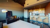 Located at 707 Prudden St in Lansing, Michigan, this single-floor loft showcases a bold industrial-chic aesthetic defined by soaring ceilings with exposed ductwork and structural beams, polished concrete floors, and an open-concept layout that seamlessly connects the living, dining, and kitchen areas. The living room is anchored by large, multi-pane factory-style windows that flood the space with natural light and offer sweeping outdoor views, creating an airy and expansive atmosphere. The kitchen is well-appointed with warm maple cabinetry, granite countertops, stainless steel appliances, and a peninsula breakfast bar accented by modern pendant lighting and contemporary bar seating — ideal for casual dining and entertaining. Rich, deep-toned accent walls add warmth and contrast against the industrial elements throughout the space. The bedroom features a distinctive wrought iron bed frame, open wardrobe storage, and a sliding barn-style door, maintaining the loft's urban character. The bathroom offers slate-tiled walls surrounding the tub and shower, polished concrete flooring, and a floating wood vanity with a vessel sink, delivering a spa-like, artisanal finish. Throughout the space, the interplay of raw materials and refined finishes creates a cohesive, sophisticated urban living environment.