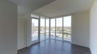 This single-floor high-rise residence offers a striking blend of modern design and expansive urban views. The standout feature of the living space is the dramatic floor-to-ceiling corner window system, composed of large glass panels framed in sleek white aluminum. These wraparound windows flood the interior with abundant natural light while providing sweeping, unobstructed panoramic views of the surrounding cityscape, neighboring high-rises, and distant hills or mountains along the horizon. A glass door within the window wall opens directly to a balcony, seamlessly connecting the interior to the outdoors. The flooring throughout is rich, dark-toned hardwood, lending warmth and contrast to the bright, airy atmosphere created by the white walls and ceiling. The clean, minimalist architectural lines are consistent throughout, with smooth white surfaces and recessed doors featuring louvered panels that add a subtle textural detail. The open layout and corner glass design create a sense of generous, light-filled space that feels both sophisticated and functional. The elevated position of the unit enhances the sense of openness, making the panoramic city views a defining and ever-present element of the living experience.
