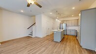 This two-story home showcases a clean, contemporary design with light-toned luxury vinyl plank flooring running consistently throughout both levels, complemented by crisp white walls and bright recessed lighting. The main floor is anchored by an open-concept layout where the kitchen flows seamlessly into the living area. The kitchen is a standout feature, equipped with gray shaker-style cabinetry, white quartz countertops, a full suite of stainless steel appliances, and a central island with a single-basin sink and elegant glass pendant lighting overhead. The living area benefits from abundant natural light provided by multiple windows with horizontal blinds, and a ceiling fan adds practical comfort. A door from the living area provides convenient exterior access. A staircase with a wooden handrail connects the main floor to the upper level. Upstairs, the bedrooms continue the cohesive aesthetic with matching flooring, vaulted ceilings, ceiling fans, and windows that draw in natural light. The primary bathroom is notably well-appointed, featuring a dual-sink vanity with quartz countertops, gray cabinetry, a large mirror spanning the full width, and a separate shower. Throughout, the home maintains a bright, move-in-ready atmosphere with consistent finishes.