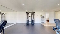 Located in Santa Clarita, CA, this dedicated fitness center offers a well-appointed exercise space designed to support a comprehensive workout routine. The single-floor layout provides a generous, open floor plan with room to move freely between equipment zones.

The space features durable dark rubber flooring throughout the main exercise area, which transitions to light hardwood-style flooring near the entry and an adjacent area with shelving. Recessed lighting illuminates the room evenly, while multiple large windows along the walls allow for ample natural light and views of the surrounding greenery, creating a bright and energizing atmosphere.

The cardio section is equipped with treadmills, elliptical trainers, and a stationary bike, each featuring individual display screens for an interactive workout experience. A full cable cross machine anchors the strength training area, complemented by a dumbbell rack with a variety of weights and an adjustable workout bench. A full-length mirror spans one wall, enhancing the sense of space and allowing users to monitor their form during exercise.

The clean, neutral-toned walls and uncluttered layout contribute to an organized and motivating environment. An exit door provides direct outdoor access, adding convenience and ventilation options. This fitness center is a well-rounded facility suited for both cardio and strength training needs.