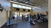 This fitness center is a thoughtfully designed, single-floor facility built to support a full range of workout routines. The space features an open floor plan with a durable, dark rubberized floor that accommodates a comprehensive selection of exercise equipment, including stationary bikes, ellipticals, cable machines, and free weights. Large floor-to-ceiling windows along one wall flood the interior with abundant natural light while offering views of the outdoor courtyard, creating an energizing atmosphere. Multiple flat-screen televisions are mounted at eye level, providing entertainment during workouts. The exposed ductwork and industrial-style ceiling add a modern, utilitarian aesthetic that complements the active environment. A standout decorative element is the dynamic wall mural depicting silhouetted figures in motion, reinforcing the space's fitness-focused character. The high ceilings contribute to an open, airy feel, preventing the space from feeling confined despite its dense equipment layout. Overhead fluorescent lighting ensures the entire facility remains well-lit. Overall, the fitness center strikes a balance between functionality and visual appeal, making it a welcoming and well-appointed environment for exercise.