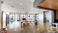 Located at 614 Rigsbee Ave in Durham, NC, this contemporary amenity space offers a well-appointed, open-concept layout with a refined modern aesthetic. Polished concrete floors flow throughout the main areas, reflecting abundant natural light that pours in through floor-to-ceiling black-framed windows and glass doors. The spacious living room is anchored by a striking textured stone accent wall housing a wall-mounted TV, complemented by a colorful area rug and versatile seating arrangements. A vertical wood slat partition elegantly divides zones while maintaining an open, airy feel. The dining and lounge area features a long communal table with contemporary bar stools beneath globe pendant lights, ideal for gathering or co-working. A cozy, boutique-style private room with a bold teal accent wall, warm wood paneling, and a round table offers an intimate setting for meetings or focused work. The expansive balcony, covered by an exposed wood-beam pergola, extends the living space outdoors with views of the surrounding Durham streetscape, furnished with bistro-height tables and lounge seating. Throughout the space, thoughtful design details — including curated artwork, mixed materials, and layered lighting — combine to create a vibrant, welcoming environment.