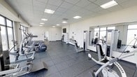 This modern fitness center is thoughtfully designed to provide a complete and comfortable workout environment. The space is finished with durable rubber tile flooring throughout, offering both safety and functionality for a wide range of exercises. A suspended drop ceiling with recessed panel lighting ensures the room is evenly and brightly illuminated, contributing to an energizing atmosphere.

The facility is generously stocked with an impressive selection of exercise equipment. Cardio machines, including treadmills, ellipticals, and stationary bikes, are arranged to allow ample space between each unit for a comfortable workout experience. A dedicated free weights area features a standing dumbbell rack stocked with multiple weight sets, catering to strength training needs. Additional resistance and weight machines round out the equipment offerings, providing options for a full-body workout.

A large wall-mounted mirror along one side of the room enhances the sense of space while allowing users to monitor their form. A flat-screen television is mounted on the wall, adding an entertainment element during workouts. Natural light filters in through a window, complementing the overhead lighting. Access doors are conveniently positioned within the space, and the clean white walls maintain a crisp, professional aesthetic throughout.