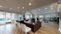 This thoughtfully designed single-floor commercial space functions as a leasing office or community clubhouse, offering a warm and professional atmosphere throughout. The open floor plan is anchored by rich hardwood flooring, neutral gray walls, and crisp white crown molding that lend a polished, cohesive aesthetic.

At the entry, a reception area welcomes visitors with dark espresso-finished desks paired with white upholstered chairs, all bathed in natural light streaming through full-length glass French doors. White structural columns define the space without interrupting sightlines, preserving the airy, connected feel of the interior.

The lounge area is a standout feature, boasting soaring vaulted ceilings trimmed in white, a dramatic floor-to-ceiling stone fireplace topped with a classic white mantel, and a wall-mounted flat-screen television flanked by arched windows that flood the room with natural light. Comfortable seating — including a neutral sectional, teal wingback chairs, and a patterned area rug — creates an inviting gathering space.

Adjacent to the lounge, a dedicated workstation area accommodates staff with dark-toned desks and ergonomic seating. A quieter lounge nook with a cozy sofa and a large window provides a more private retreat. The space also features recessed lighting throughout, adding warmth and functionality across all zones.