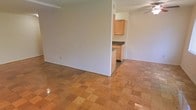 This single-floor space offers a straightforward and functional layout designed for comfortable everyday living. The main living area is finished with classic parquet-style wood flooring that adds warmth and character to the space. Neutral white walls keep the interior bright and versatile, providing a clean backdrop that complements a variety of décor styles. A ceiling fan is installed overhead, contributing to year-round comfort and air circulation throughout the open room.

Adjacent to the living area, the kitchen is partially open to the main space, separated by a partial dividing wall that maintains a sense of connection between the two areas. The kitchen features light oak wood cabinetry that provides ample storage, paired with dark countertops that offer a subtle contrast. The layout appears practical and efficient, making good use of the available space for meal preparation and storage.

Overall, the space presents a clean, no-frills interior that prioritizes functionality. The consistent flooring, open flow between living and kitchen areas, and simple finishes make it an adaptable space suitable for a range of uses and personal styles.
