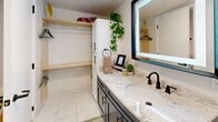 Located at 609 S Thornton Ave in Dalton, Georgia, this thoughtfully designed bathroom and closet combination delivers a clean, modern aesthetic with carefully selected finishes throughout. The space is anchored by a stylish single-sink vanity topped with a speckled granite countertop and fitted with a matte black faucet, adding a contemporary contrast to the light stone surface. Above the vanity, a backlit LED mirror with a bold dark frame provides functional illumination while enhancing the room's modern character. The glass-enclosed shower features crisp white subway tile walls, a built-in niche shelf, and matte black hardware including a towel bar, maintaining a cohesive design language across the space. Marble-look porcelain tile flooring extends throughout, contributing to a bright and polished atmosphere. An integrated closet area with dual hanging rods, open wood shelving, and a tall white storage cabinet with matte black pulls offers organized and accessible storage. The white cabinetry and neutral wall tones create a light-filled, airy environment. Every detail reflects a commitment to both style and practicality, making this a refined and functional space.