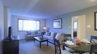 This single-story space centers around a combined living and dining area that feels open and cohesive in its layout. The living area is anchored by plush wall-to-wall carpeting and features soft blue-toned walls that lend a calm, relaxed atmosphere throughout. Two windows allow generous natural light to filter in, brightening the space and creating an airy feel. The seating arrangement accommodates both lounging and conversation, with sufficient room for a sofa, armchair, and a rectangular coffee table. A framed piece of wall art adds a decorative focal point above the sofa. Adjacent to the living area, a dining set with dark wood chairs is partially visible, suggesting a seamless transition between living and dining functions. A doorway leads to what appears to be a bedroom, offering a glimpse of additional carpeted space beyond. The overall ambiance is comfortable and functional, with a consistent color palette of cool blues and neutral tones tying the space together. The layout makes efficient use of the available floor plan while maintaining a sense of openness.