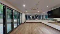 This single-floor fitness and wellness facility is designed with both function and ambiance in mind. The centerpiece is a spacious studio with wide-plank hardwood flooring, floor-to-ceiling glass panel walls lined with ballet barres, and a large wall-mounted display screen for guided sessions. Recessed lighting and a ceiling-mounted HVAC unit ensure a comfortable environment year-round. Expansive folding glass doors open directly to a private outdoor courtyard, allowing natural light and views of towering bamboo to fill the studio interior. Equipment such as yoga mats and blocks are neatly stored in wall-mounted racks, keeping the space clean and organized. Adjacent to the studio is a moody, dark-toned lounge area featuring a built-in bench, wall-mounted television, and a dramatic nature-inspired photo mural, providing a relaxing retreat before or after activity. A dedicated golf simulator room, finished in deep charcoal tones with full swing simulator technology and turf putting green, adds a recreational dimension to the facility. The outdoor courtyard is a highlight — enclosed by concrete planters overflowing with lush bamboo, it features tiled flooring, stylish lounge seating, and subtle ground-level lighting, creating a tranquil, Zen-inspired retreat.