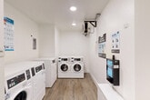 This shared laundry facility at 6101 Richmond Avenue in Houston, Texas offers a clean, efficiently organized space designed for convenient, everyday use. The room features a bright, modern aesthetic with crisp white walls, white-painted trim, and wood-look flooring that adds a touch of warmth throughout.

Multiple commercial-grade front-loading washing machines and dryers are arranged along the walls, providing ample capacity for simultaneous loads. The machines feature digital control panels, offering a range of cycle options to suit various laundry needs. A wall-mounted payment kiosk is conveniently positioned within the room, streamlining the payment process with easy-to-follow posted instructions nearby.

Additional amenities include a wall-mounted air conditioning unit for year-round climate comfort, a built-in hand dryer, and a small white storage cabinet that provides supplementary counter space. Recessed lighting keeps the room well-illuminated, while a security camera ensures the safety of users. Two access doors offer easy entry and exit. The overall layout is thoughtfully arranged to allow users to move freely and comfortably while managing their laundry, making this a practical and well-appointed shared amenity.