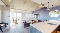Located at 1590 Elmwood Ave in Evanston, Illinois, this thoughtfully designed unit offers a seamless open-concept layout that integrates the kitchen and living areas into one cohesive, functional space. The living room is anchored by a bold periwinkle accent wall, wood-plank-style flooring, and a striking wood-paneled ceiling that adds warmth and texture. Large windows and a sliding glass door flood the space with abundant natural light while framing impressive city skyline views. A private balcony accessed from the living area provides an intimate outdoor retreat. The modern kitchen is a standout feature, showcasing dark wood cabinetry, a blue subway tile backsplash, stainless steel appliances including a gas range and full-size refrigerator, and a generous island with a white quartz countertop and bar seating for multiple guests. Pendant lighting with metallic gold accents enhances the kitchen's contemporary aesthetic. Residents also enjoy access to a spacious rooftop terrace complete with lounge seating, a fire pit, planters with lush greenery, a grill station, and panoramic views of the surrounding Evanston cityscape — ideal for relaxing or entertaining under the open sky.