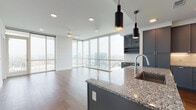 This two-bedroom, one-and-a-half-bath residence offers a refined urban living experience defined by its striking floor-to-ceiling glass walls that frame panoramic city skyline views throughout the main living areas. Rich hardwood flooring runs through the open-concept living and dining space, which flows seamlessly into a well-appointed kitchen. The kitchen is outfitted with slate-gray shaker cabinetry, a white subway tile backsplash, stainless steel appliances, granite countertops, and a generous center island with pendant lighting overhead. A built-in wine rack and additional cabinetry provide ample storage. The primary bedroom features expansive glazing with direct balcony access, flooding the room with natural light and city views, and is complemented by a spacious walk-in closet with built-in shelving. The primary bath offers a dual-sink granite vanity, tile flooring, and a soaking tub with full tile surround. A second carpeted bedroom provides a quiet retreat, while the half bath features a granite vanity and clean tile flooring. Throughout, the design balances contemporary finishes with a bright, airy ambiance.