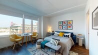 This contemporary single-floor unit is thoughtfully designed to maximize both comfort and functionality. The main living and sleeping area is the heart of the space, characterized by clean white walls, dark hardwood flooring, and large windows that flood the room with natural light and frame sweeping urban views of the surrounding cityscape. A cozy seating nook positioned by the windows features a bistro-style table and vibrant yellow accent chairs, creating an inviting area for relaxing or working. A gallery wall of six colorful framed artworks adds a lively, creative touch to the sleeping area, complementing the contemporary aesthetic throughout.

The kitchen is a standout feature, showcasing bold navy lower cabinetry paired with light wood upper cabinets and sleek gray countertops. An elongated hexagonal tile backsplash adds a modern geometric element, while stainless steel appliances — including a dishwasher, range, and built-in microwave — provide full functionality in a compact, efficient layout.

The bathroom continues the geometric design theme with striking large-format hexagonal wall tiles surrounding a full soaking tub and shower combination. Warm-toned floor tiles and recessed lighting contribute to the clean, spa-like atmosphere of the room. Overall, the space presents a cohesive, modern aesthetic enhanced by bold accent colors, thoughtful storage solutions, and an abundance of natural light.