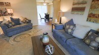 This single-story space features an open-concept layout that seamlessly connects the living and dining areas, creating a functional and welcoming environment. The living area is anchored by plush, cushioned sofas arranged around a wooden coffee table, accented by decorative elements including ceramic vases and sculptural accents that add visual interest. A patterned area rug defines the seating zone, lending warmth and texture to the carpeted floor. Ambient lighting is provided by a stylish table lamp, complementing the natural light that filters in from a sliding glass door at the far end of the space. The dining area, visible from the living room, features a ceiling fan with integrated lighting and is furnished with a dining table and chairs. Vertical blinds on the sliding door allow for adjustable light control while maintaining a connection to the outdoors. The warm, neutral-toned walls throughout contribute to a cohesive and calming atmosphere. The overall layout is practical and well-proportioned, offering a comfortable flow between the social spaces of the home.