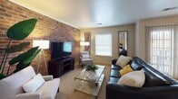 This thoughtfully designed single-floor residence offers a comfortable and inviting layout across several distinct living areas. The living room serves as the centerpiece of the home, highlighted by a dramatic full-height exposed brick accent wall that adds warmth and character to the space. Plush carpeting, natural light from white-blinded windows, and ample room for seating create an ideal environment for relaxing or entertaining. The kitchen features white cabinetry, laminate countertops, a stainless steel sink, and a full suite of appliances including an electric range and dishwasher, all set on durable tile flooring. The primary bedroom is a serene retreat with soft carpet, dual windows for natural light, a ceiling fan, and rich cherry wood furniture. The second bedroom mirrors the cozy feel with its own ceiling fan, carpeted floors, and natural light. Both bathrooms are clean and functional — one featuring a tiled floor and pedestal-style sink, and the other a full tub-shower combination with a wood-toned vanity. Throughout the residence, neutral tones and warm lighting contribute to a calm and cohesive ambiance.
