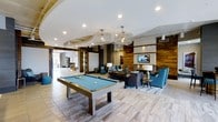 Located at 111 Cowan St in Wilmington, NC, this single-floor amenity space offers a thoughtfully designed communal environment that blends modern aesthetics with comfortable living. The open-plan layout creates a seamless flow between activity and relaxation zones, anchored by a striking pool table positioned beneath two statement wire-frame chandeliers that cast warm, ambient light throughout the room.

The lounge area is centered around a classic fireplace framed by a wood accent wall, which adds texture and warmth to the contemporary interior. A mounted flat-screen television above the fireplace enhances the space's entertainment appeal. Plush seating, including rich blue armchairs and a charcoal sofa, is arranged to encourage conversation and relaxation around the hearth.

Light wood-toned flooring runs throughout, reflecting natural light and contributing to the bright, airy atmosphere. Exposed wooden ceiling beams add an architectural character that contrasts beautifully with the sleek white ceilings and recessed lighting above. A barn-style sliding door and open sightlines to adjacent areas suggest a spacious, interconnected layout designed for community engagement. The overall ambiance is sophisticated yet inviting, making this a well-appointed gathering space for residents.