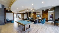 Located at 111 Cowan St in Wilmington, NC, this single-floor amenity space offers a thoughtfully designed communal environment that blends modern aesthetics with comfortable living. The open-plan layout creates a seamless flow between activity and relaxation zones, anchored by a striking pool table positioned beneath two statement wire-frame chandeliers that cast warm, ambient light throughout the room.

The lounge area is centered around a classic fireplace framed by a wood accent wall, which adds texture and warmth to the contemporary interior. A mounted flat-screen television above the fireplace enhances the space's entertainment appeal. Plush seating, including rich blue armchairs and a charcoal sofa, is arranged to encourage conversation and relaxation around the hearth.

Light wood-toned flooring runs throughout, reflecting natural light and contributing to the bright, airy atmosphere. Exposed wooden ceiling beams add an architectural character that contrasts beautifully with the sleek white ceilings and recessed lighting above. A barn-style sliding door and open sightlines to adjacent areas suggest a spacious, interconnected layout designed for community engagement. The overall ambiance is sophisticated yet inviting, making this a well-appointed gathering space for residents.