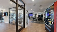 This fitness center in Plano, Texas, offers a thoughtfully organized, single-floor layout designed to accommodate a wide range of workout routines. The space is defined by its striking floor-to-ceiling glass wall panels and bold black-framed glass entry doors, which flood the interior with natural light and provide open sightlines throughout.

The strength training area features a power rack with weight plates, a dedicated dumbbell rack with a full range of free weights, multiple selectorized cable machines, and weight benches positioned across durable hardwood-style flooring. A wall-mounted flat-screen television and a deep navy accent wall anchor the free weights zone, creating a distinct, focused atmosphere. Full-length mirrors are strategically placed throughout, enhancing the sense of depth and supporting proper form during exercise.

The cardio zone, positioned along the glass perimeter, houses treadmills, a stair climber, ellipticals, and stationary bikes, all benefiting from natural light and outdoor views. Centrally located floor mat space provides a functional area for stretching and bodyweight training. Recessed lighting and ceiling fans contribute to a clean, comfortable environment. The facility's layout ensures efficient movement between zones, supported by clear corridor access and well-placed entry and exit points.