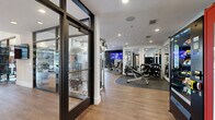 This fitness center in Plano, Texas, offers a thoughtfully organized, single-floor layout designed to accommodate a wide range of workout routines. The space is defined by its striking floor-to-ceiling glass wall panels and bold black-framed glass entry doors, which flood the interior with natural light and provide open sightlines throughout.

The strength training area features a power rack with weight plates, a dedicated dumbbell rack with a full range of free weights, multiple selectorized cable machines, and weight benches positioned across durable hardwood-style flooring. A wall-mounted flat-screen television and a deep navy accent wall anchor the free weights zone, creating a distinct, focused atmosphere. Full-length mirrors are strategically placed throughout, enhancing the sense of depth and supporting proper form during exercise.

The cardio zone, positioned along the glass perimeter, houses treadmills, a stair climber, ellipticals, and stationary bikes, all benefiting from natural light and outdoor views. Centrally located floor mat space provides a functional area for stretching and bodyweight training. Recessed lighting and ceiling fans contribute to a clean, comfortable environment. The facility's layout ensures efficient movement between zones, supported by clear corridor access and well-placed entry and exit points.