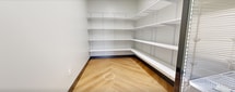 Located at 85 Livingston Ave in Roseland, NJ, this 810-square-foot, single-floor facility serves as a well-appointed mail and package management center for residents. The space is organized across three distinct areas, each designed with both function and aesthetics in mind.

The main mail lobby is a standout space, showcasing a coffered ceiling with elegant pendant chandeliers that cast warm light throughout the room. A large, centrally positioned wood-framed mailbox island houses rows of individual tenant mailboxes with a refined, light natural wood enclosure. The floor features a warm herringbone-patterned tile, adding visual texture and sophistication. A glass-enclosed package locker kiosk is also present, providing secure parcel pickup.

An adjacent corridor continues the herringbone flooring and is lined with warm wood wall paneling accented by sleek wall sconces. Additional mailbox banks line this hallway, offering ample individual mail storage capacity.

A dedicated package and cold storage room features a commercial-grade glass-door refrigeration unit for chilled deliveries, alongside a separate storage room with floor-to-ceiling adjustable white shelving and a herringbone wood-look floor. A corridor with direct exterior access ensures efficient package delivery logistics. Together, these areas form a cohesive, polished amenity space built for modern residential convenience.