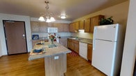This single-floor residence offers a functional and inviting layout designed for comfortable everyday living. The kitchen serves as a standout feature of the home, showcasing rich honey-toned wood cabinetry that extends to the upper walls, providing generous storage throughout. Granite-look countertops line the perimeter of the kitchen, complemented by a matching central island that doubles as a casual dining and prep space, complete with built-in electrical access. White appliances — including a refrigerator, dishwasher, range, and over-the-range microwave — lend a clean, cohesive aesthetic. A stylish multi-bulb pendant chandelier hangs above the island, adding a warm focal point to the space, while recessed and flush-mount ceiling lighting ensures the kitchen remains well-lit. Warm hardwood flooring flows throughout, adding a sense of continuity and natural warmth. The kitchen's L-shaped layout maximizes counter and cabinet space while maintaining an open, easy-to-navigate flow. The overall ambiance of the home is welcoming and practical, with warm tones and classic finishes that create a timeless residential feel suited for a variety of lifestyles.