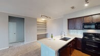 This 2-bedroom, 2-bathroom residence in Humble, TX is thoughtfully laid out across a single floor, offering a functional and comfortable living environment. The open-concept kitchen serves as the heart of the home, featuring dark espresso cabinetry, light quartz countertops, a breakfast bar island with an undermount sink and modern faucet, black appliances including a refrigerator with ice maker, electric range, and microwave, plus a subway tile backsplash and hardwood-style flooring. Track lighting illuminates both the kitchen and the adjoining built-in shelving unit, which adds practical storage and display space. The living area is carpeted and bathed in natural light from large windows and a glass-paneled door, accented by crown molding and a ceiling fan. Both bedrooms are carpeted and well-sized; the primary bedroom features dual windows with outdoor views, crown molding, and a ceiling fan, and connects to an en-suite bathroom with a granite-topped vanity, tub/shower combo, and a walk-in closet. The second bathroom mirrors the same dark cabinetry and granite vanity finishes. A dedicated laundry closet with washer/dryer connections adds everyday convenience.