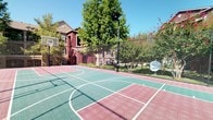 This outdoor recreational amenity space centers around a full-size multi-sport court finished in a distinctive green and burgundy tile surface, clearly marked with basketball and additional court lines. Two basketball hoops are positioned at each end of the court, and the entire playing area is enclosed by tall, heavy-duty safety netting supported by sturdy poles, ensuring safe play and containment. A concrete walkway borders the court, providing easy access from all sides.

The surrounding grounds are thoughtfully landscaped, featuring well-maintained green lawns, mature shade trees, and blooming crape myrtles that add seasonal color. Stone retaining walls line nearby pathways, filled with decorative gravel and lush shrubs, adding a polished, structured aesthetic to the outdoor environment. The natural elements create a pleasant, park-like atmosphere around the athletic space.

The backdrop includes multi-story residential buildings with classic architectural details — stone accents, stucco exteriors, balconies, and pitched rooflines — that frame the space with a sense of community. The open-air setting benefits from abundant natural light and clear sightlines to mature trees and blue skies, creating an inviting environment for active recreation throughout the day. This well-appointed sport court offers a premier outdoor amenity within a vibrant residential community.