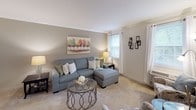 This single-floor residence offers a thoughtfully arranged layout with distinct living spaces connected by a cohesive neutral color palette and comfortable finishes. The living room is a welcoming area featuring carpet flooring, crown molding, and natural light from a large window framed by draping curtains. The space comfortably accommodates a sectional sofa and accent furnishings, creating a relaxed yet polished atmosphere.

Adjacent to the living room, the dining area continues the refined aesthetic with wainscoting panels lining the lower walls, crown molding at the ceiling, and carpet underfoot. A glass-top dining table with seating for four fits naturally within the space.

The kitchen is efficiently designed with warm oak cabinetry, dark granite-style countertops, light hardwood flooring, and a window that brings in natural light. It includes a full suite of appliances and ample upper and lower cabinet storage.

The residence features multiple bedrooms, each carpeted and bright with natural light from individual windows. The bedrooms offer generous space for full-sized furnishings and include closet access. The full bathroom features tile flooring, a tub-shower combination with tile surround, and a clean, functional layout.