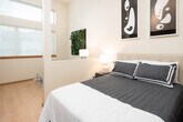 This well-appointed single-floor studio offers a smart, efficient layout that maximizes every inch of available space. The sleeping area is defined by a partial structural column and low partition wall, creating a sense of separation from the living area while maintaining an open, airy feel. The bedroom is warmly lit by a pair of bedside lamps and decorated with bold abstract artwork, complemented by light hardwood flooring that runs throughout the entire unit. Large windows fitted with horizontal blinds allow generous natural light to fill the space, offering views of lush greenery outside.

The living area flows naturally from the sleeping zone, featuring a comfortable seating arrangement atop a textured area rug, with tall accent lighting adding warmth to the neutral-toned walls. A compact kitchenette lines one wall, outfitted with rich dark wood cabinetry, open shelving, a stainless steel sink, and full appliances including a refrigerator and range — providing full functionality in a streamlined footprint.

The bathroom is clean and practical, featuring a single vanity with a wide mirror, grey countertop, and a stacked washer/dryer unit conveniently integrated into the same space. The overall ambiance is modern and calm, with a cohesive neutral palette and contemporary finishes throughout.