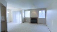 This single-floor residential space offers a clean, neutral aesthetic throughout. The living area serves as the centerpiece of the home, showcasing wall-to-wall plush carpeting that adds warmth and comfort underfoot. A classic fireplace with a tiled surround and raised hearth anchors one wall, providing both a functional heating source and a natural focal point for the room. The space is bathed in natural light, courtesy of multiple windows fitted with horizontal blinds, as well as a large sliding glass door dressed with vertical blinds that likely leads to an outdoor area. The walls are finished in a soft, warm neutral tone that complements the light-colored carpet, creating a cohesive and inviting atmosphere. Ceiling-mounted lighting fixtures provide additional illumination throughout the space. The overall design is understated and versatile, offering a blank canvas that adapts easily to a variety of interior styles. The open layout and abundant natural light contribute to an airy, spacious feel that makes the living area both functional and welcoming.