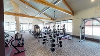 This fitness center is a thoughtfully designed workout facility that combines functional equipment with striking architectural character. The space is defined by its dramatic vaulted ceiling adorned with exposed natural wood beams, which lend a warm, lodge-inspired aesthetic to an otherwise modern gym environment. Large windows line the walls, flooding the interior with natural light and providing open views to the outside, creating an energizing atmosphere for workouts.

The facility is outfitted with a comprehensive selection of equipment to accommodate a wide range of fitness routines. A full dumbbell rack with multiple weight increments anchors the free weights area, accompanied by weight benches and selectorized strength machines for targeted resistance training. The cardio section features a row of elliptical trainers and treadmills positioned near the windows, allowing users to exercise while enjoying natural light and outdoor views.

Ceiling fans are mounted throughout the space to maintain comfortable airflow, while patterned carpeting and rubberized flooring define distinct workout zones. The overall ambiance is spacious, bright, and welcoming, offering a well-rounded fitness environment suited for both strength training and cardiovascular exercise.